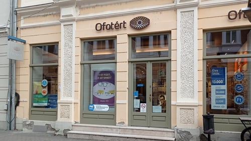 Ofotért optika utcai homlokzata Baján, az Eötvös József utcában, üvegportálokkal és kirakati plakátokkal.