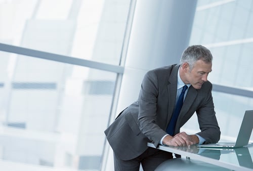 A man in a gray suit leans over a desk, focused on a laptop in a modern office with large windows. The scene conveys a professional and concentrated atmosphere.