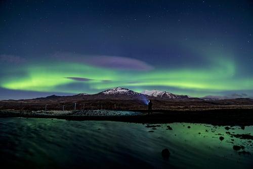 Person viewing aurora borealis over snow-capped mountains and stream in Arctic landscape at night.