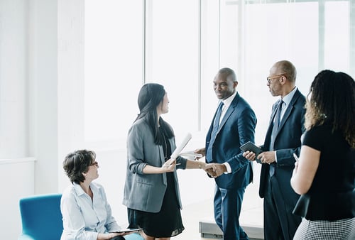 A group of diverse professionals in a bright office engage in conversation. Two men in suits shake hands, while a woman seated with a tablet looks on. The mood is collaborative.
