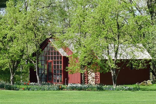 Red barn-style building with large glass-paned gable entrance housing William Pitt Sotheby's International Realty in Millbrook, New York, surrounded by spring foliage and landscaped lawn with flowering bulbs.