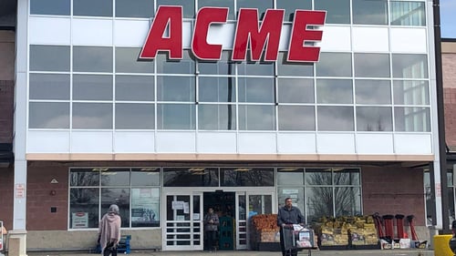 Man with full shopping cart walking out of Aceme grocery store while woman with shawl walks in