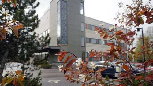 Kuva kolmikerroksisesta rakennuksesta, jonka etualalla parkkipaikkoja ja ruskansävyisiä pensaita ja puita.