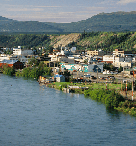 A riverside town with industrial buildings and vibrant greenery, set against distant, forested mountains under a clear sky, conveys tranquility.