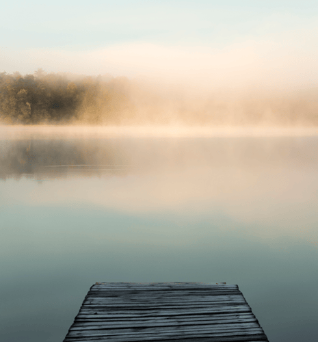 A wooden dock extends over a calm lake enveloped in light mist. The far shore features autumn trees with golden leaves reflected in the water, creating a tranquil scene.
