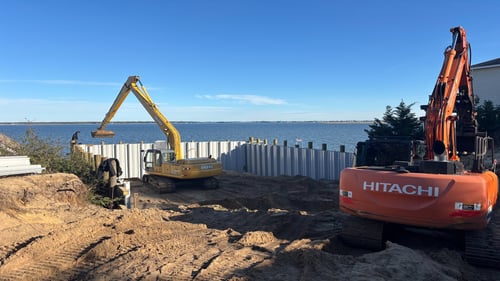 Construction equipment working along a waterfront site, with a yellow John Deere excavator and an orange Hitachi excavator placing sheet piling beside the water under a clear blue sky.