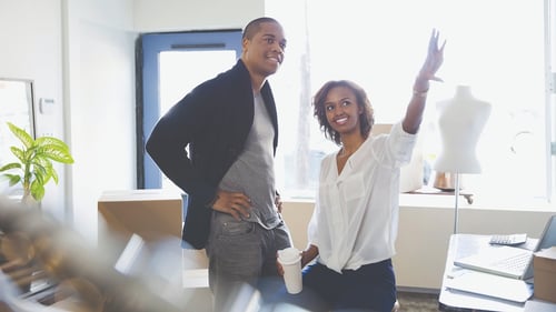 Two people in a bright space discuss ideas. A woman, smiling, gestures upward with enthusiasm, holding a coffee cup. A man stands beside her, listening intently.