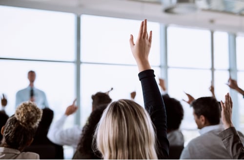 Audience members raising hands in a bright conference room. A person in front, blurred, speaks at a podium. The mood is engaged and interactive.