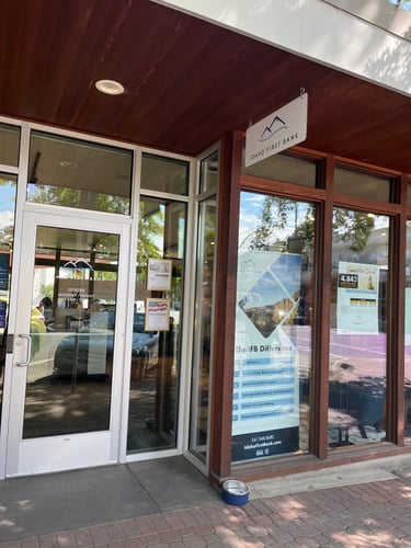 The entrance of an "Idaho First Bank" branch. A glass door shows the bank's hours, and a large poster inside a window advertises "The IFB Difference." A hanging sign with the bank's logo is visible under a wooden overhang. A small dog bowl is on the ground near the window.