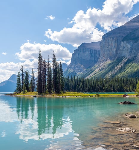 A serene lake scene with turquoise water reflecting tall evergreen trees and rocky mountains. Fluffy clouds scatter across the blue sky, evoking tranquility.