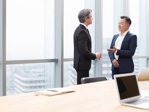 Two businessmen in suits engage in conversation in a bright office with large windows overlooking a cityscape. A laptop rests on a wooden table.
