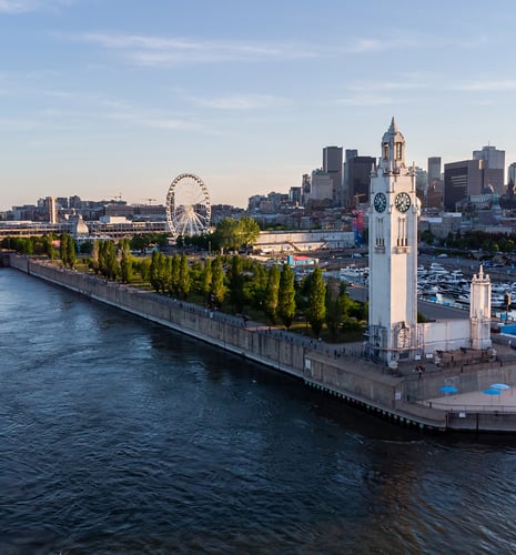 A scenic view of a city waterfront at sunset, featuring a prominent clock tower and lush green trees. A ferris wheel and city skyline are visible in the background.