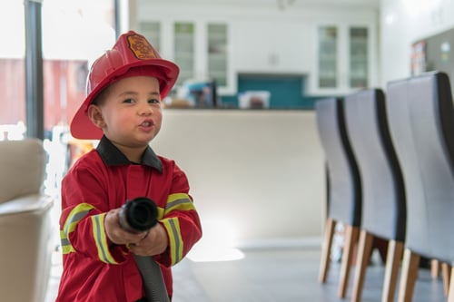 Kid points a hose to the camera while wearing a fireman costume