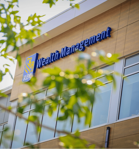Building facade with "RBC Wealth Management" in blue letters above a logo, viewed through green leaves, giving a professional and serene atmosphere.