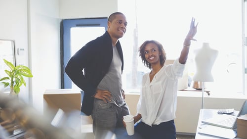 Two people in a bright space discuss ideas. A woman, smiling, gestures upward with enthusiasm, holding a coffee cup. A man stands beside her, listening intently.