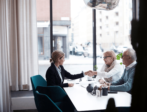 A woman in a business suit shakes hands with an older woman at a table in a modern office. An older man watches, and large windows show a city street.