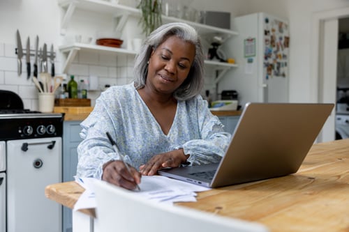 Woman writing on paper sitting at kitchen table with laptop open