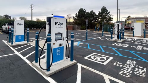 Several EVgo fast-charging units installed in marked parking spaces at a public charging station, with EVgo logos on the chargers, protective bollards around each unit