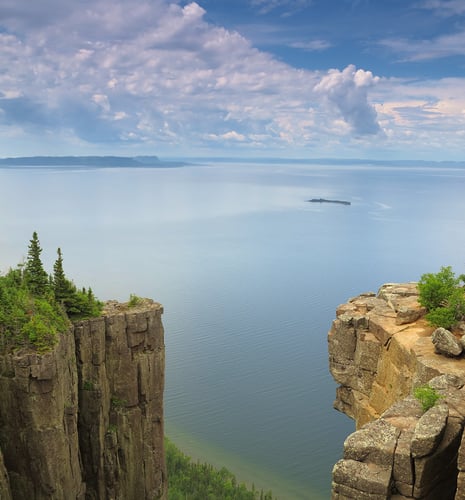 Expansive lake view framed by towering cliffs with sparse greenery. The serene sky is partly cloudy, creating a calm, tranquil atmosphere.