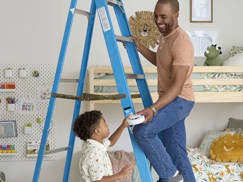 A father is accepting a smoke alarm from his son as he steps on a ladder in a child's bedroom.