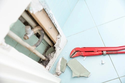 Exposed bathroom pipes behind broken drywall with a red pipe wrench.