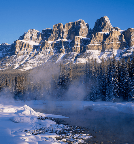 Snow-covered mountains and evergreen trees with rising mist under a clear blue sky create a serene winter landscape, conveying tranquility and majesty.