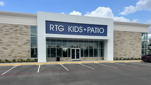 Exterior of the South Austin RTG Kids + Patio store with a modern facade, stone accents, large glass windows, and a blue sign above the entrance on a sunny day.