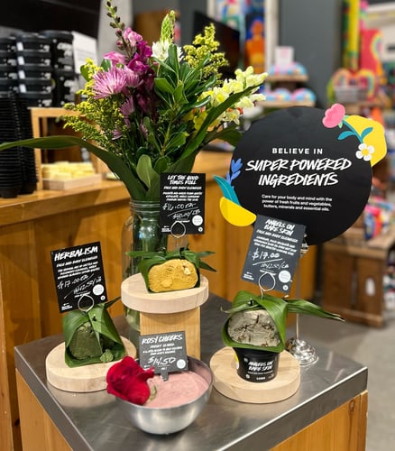 Unpackaged fresh facial cleaners wrapped in leaves, and a metal bowl containing a fresh face mask garnished with a rose. Behind them is a vase full of colourful flowers. A sign next to them reads Believe In Super Powered Ingredients. The interior of the shop is visible in the background.