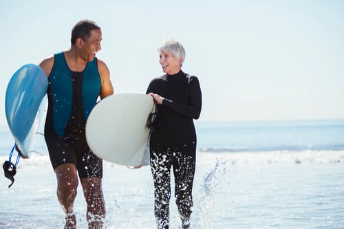 A man and woman, both in wetsuits, joyfully walk through shallow ocean water, holding surfboards and smiling, with a bright blue sky overhead.