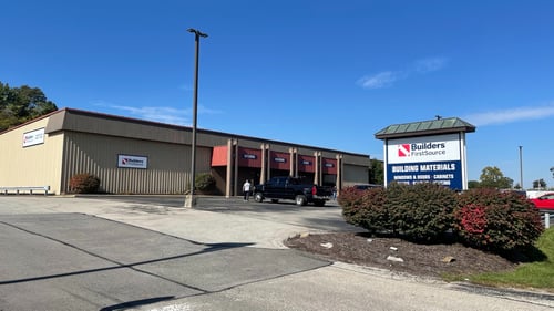 Wide view of a Builders FirstSource building materials location with a branded roadside sign, parking area, landscaping, and showroom building beneath a clear blue sky.