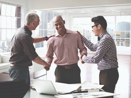 Three colleagues in a bright office celebrate with smiles and handshakes by a desk with a laptop and papers, conveying teamwork and success.