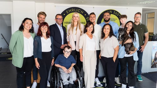 Gruppenfoto von dreizehn Personen in einem Büro. Eine Person sitzt im Rollstuhl, eine andere hält einen kleinen Hund im Arm. Die Gruppe wirkt freundlich und divers, mit Personen unterschiedlichen Alters und Erscheinungsbildes. Im Hintergrund ein Firmenlogo mit den Worten „EHRLICH“ und „VERANTWORTUNG“.