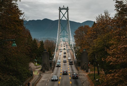 Cars drive across a suspension bridge surrounded by lush autumn foliage, with distant mountains under a cloudy sky, creating a tranquil, scenic view.