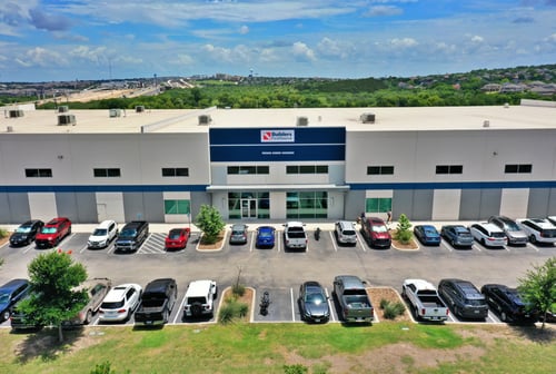 Aerial front view of the Builders FirstSource Premium Window Showroom in San Antonio, featuring a large blue-and-white commercial building with BFS signage, a covered entrance, expansive windows, and a full parking lot. Green space, trees, and a highway are visible in the background under a partly cloudy sky.