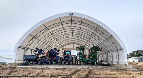Large farm machinery, including a tractor and tillage equipment, stored inside an open-ended Pilot Buildings arched truss structure.