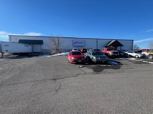 Front view of the Builders FirstSource Montrose location, featuring a metal building with the company logo displayed above the entrance. Several vehicles are parked in front of the building on a clear day with patches of snow on the ground.