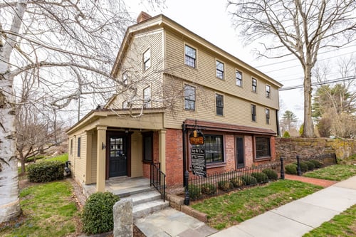 Tan three-story building with brick first floor and covered entrance housing William Pitt Sotheby's International Realty in Glastonbury, Connecticut, featuring stone wall and historic architecture.