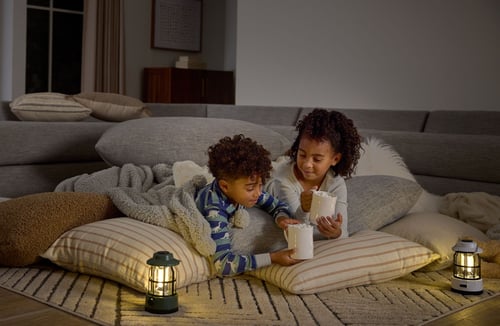 Two kids sip hot cocoa while laying on the floor in their living room. The area is lit with lanterns.