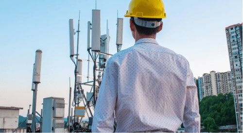 A person in a yellow hard hat and white shirt stands on a rooftop, looking at a cluster of communication antennas at sunset, with city buildings nearby.