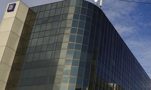 Modern office building with reflective glass facade and a logo in the corner, set against a blue, partly cloudy sky, conveying a corporate atmosphere.