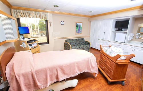 The labor and delivery room with a bed, pull-out sofa, storage cabinets, TV and bassinet.