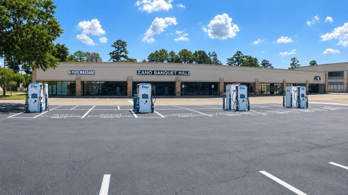 Several EVgo fast charging units installed in marked parking spaces at a public charging station, with EVgo logos on the chargers, protective bollards around each unit. CCS chargers featured in the image. A restaurant is visible in the background.