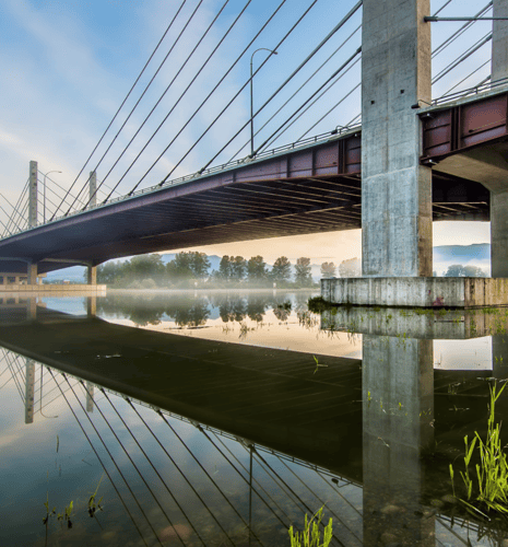 Cable-stayed bridge reflected in a calm river at sunrise. Fog lingers near distant trees, creating a serene, peaceful atmosphere.