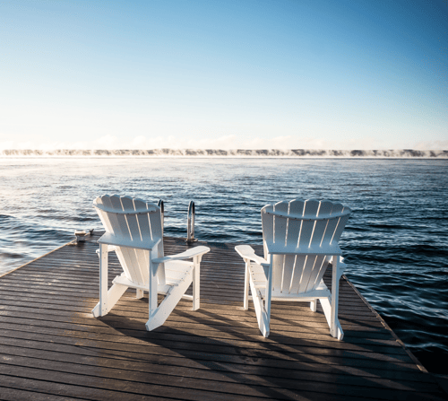 Two white Adirondack chairs on a wooden dock facing a calm ocean under a clear blue sky, evoking a peaceful and serene atmosphere.