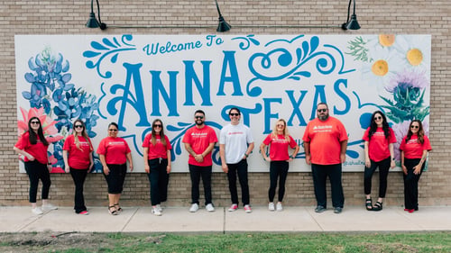 Nine Andy Torres State Farm team members stand across a sidewalk in front of a large Anna, Texas mural painted with blue lettering and flowers.