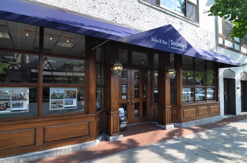 White brick building with dark wood storefront and blue awning housing Julia B. Fee Sotheby's International Realty in Bronxville, New York, featuring property listing displays in windows.