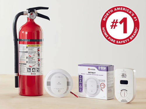 A red fire extinguisher stands upright on a wooden surface, flanked by white smoke/carbon monoxide detectors and their boxed packaging, against a pale wall bearing a circular "#1" safety-brand badge.