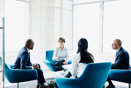 A group of four professionals sits in blue chairs in a modern office with large windows. They are engaged in a serious discussion, fostering a collaborative atmosphere.