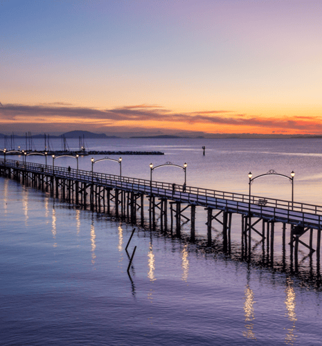 A long pier extends over calm water at sunset, with warm lights reflecting on the surface. The sky is a gradient of purple and orange hues, creating a serene atmosphere.