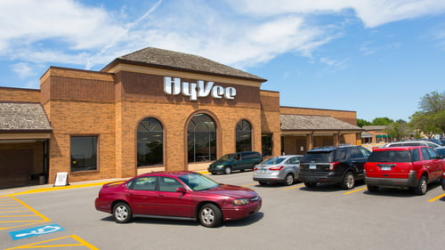 Red car passing parked cars in front of Hyvee at Westchester Square shopping center on a clear day